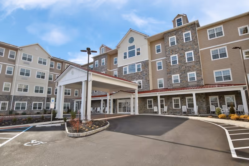 Exterior view of a multi-story senior living facility building with a covered entrance driveway, several windows, stone and beige siding, and a clear blue sky above. There are white chairs along the front porch and a handicapped parking space visible in the foreground.
