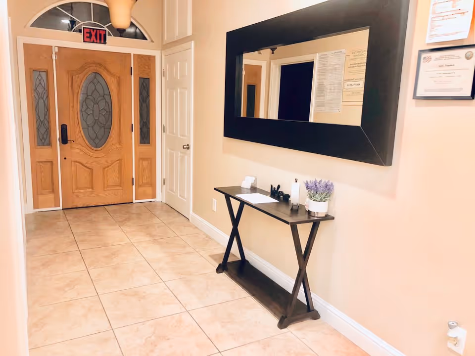 Entrance hallway of a residential care home with a wooden front door featuring decorative glass panels. A large rectangular mirror with a dark frame hangs on the right wall above a narrow wooden console table holding a small potted plant, papers, and office supplies. The floor is tiled with light-colored tiles, and an exit sign is visible above the door.
