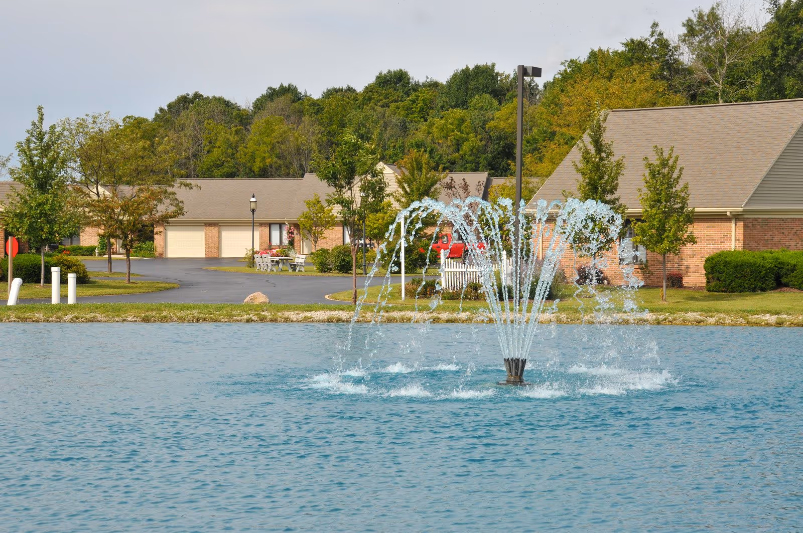 A decorative fountain sprays water in a pond with single-story brick senior living buildings, trees, and a driveway in the background.