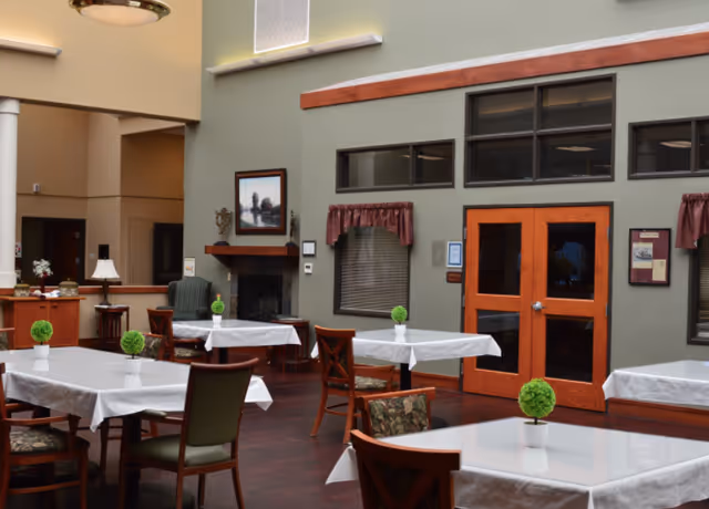 A spacious dining room with several tables covered in white tablecloths, each decorated with a small green potted plant. The room features wooden chairs with patterned cushions, a fireplace with a framed painting above it, and large windows with maroon curtains. The walls are painted in neutral tones, and there are double wooden doors with glass panels.