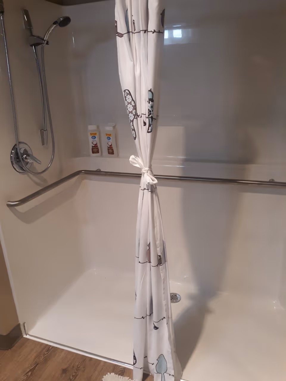 A shower area with a handheld showerhead mounted on the wall, a white shower curtain with a nature-themed pattern tied in the middle, and two bottles of shampoo or body wash placed on a built-in shelf. There is a horizontal grab bar along the back wall and wood-style flooring outside the shower.