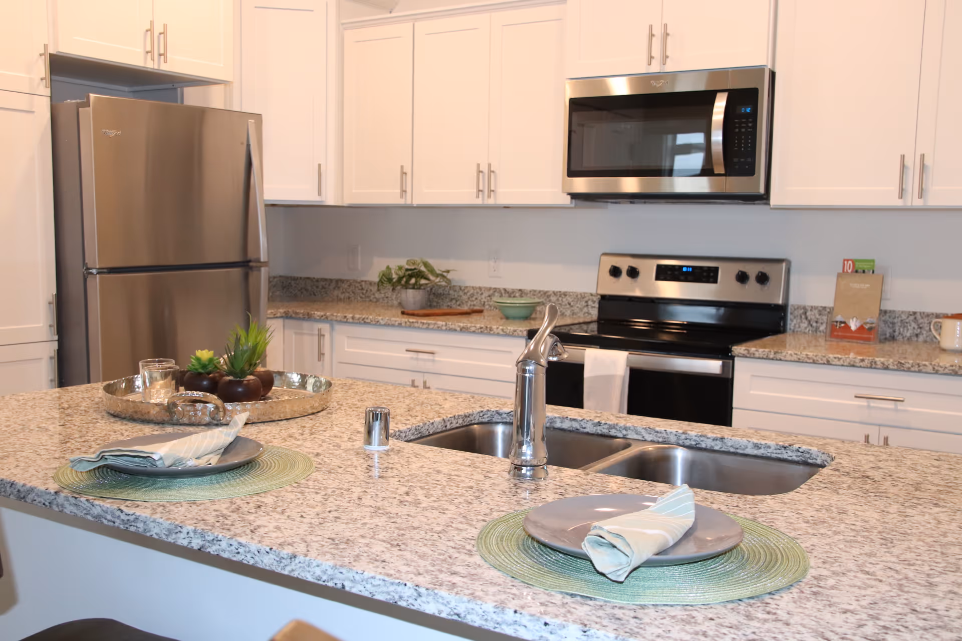 Bright modern kitchen featuring a granite island with a double sink, stainless steel refrigerator and oven, and two place settings.