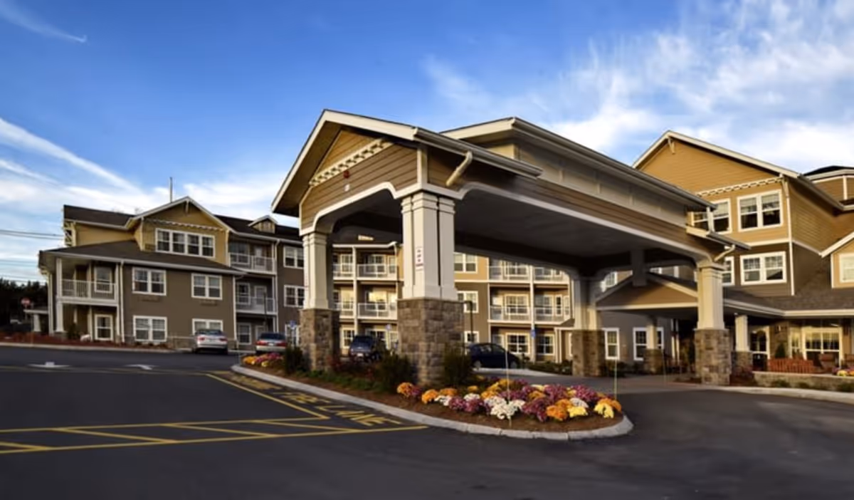 Exterior view of Fairview Estates Gracious Retirement Living building with a covered entrance supported by stone and white pillars, landscaped flower beds, parked cars, and a clear blue sky.