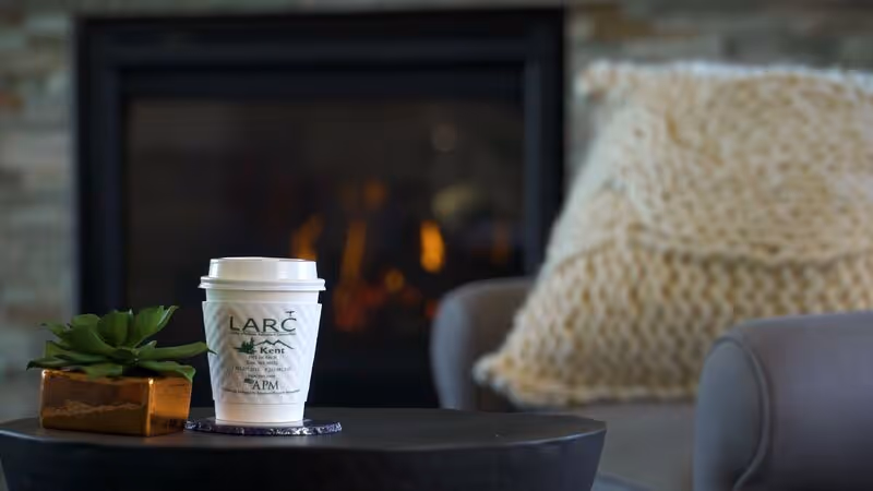 A close-up of a white disposable coffee cup with the LARC at Kent logo on it, placed on a dark round table next to a small potted succulent plant. In the background, there is a cozy living room setting with a knitted cushion on a gray armchair and a fireplace with a warm fire burning.