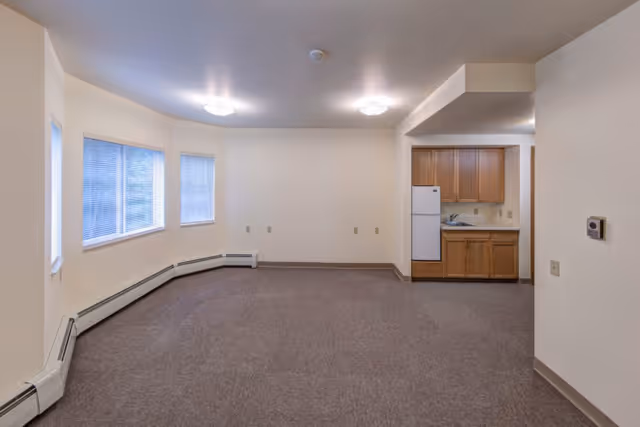 Empty room with beige walls and carpeted floor, featuring three windows with blinds on the left side and a small kitchen area with wooden cabinets, a refrigerator, and a sink on the right side.