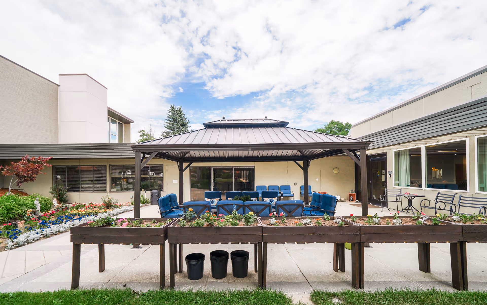 Courtyard with a covered patio of blue chairs, raised wooden planter beds and surrounding flowerbeds.