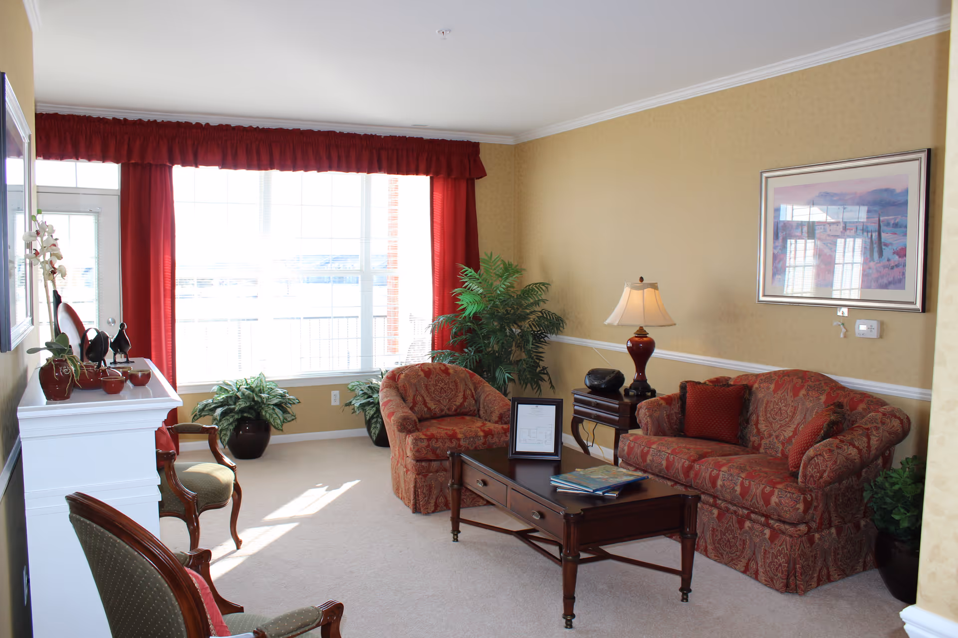 A cozy living room with a large window covered by red curtains letting in natural light. The room features a patterned red sofa and matching armchair, a wooden coffee table with books and a framed picture, a side table with a lamp, and several green potted plants. The walls are painted beige with a framed landscape painting hanging above the sofa.