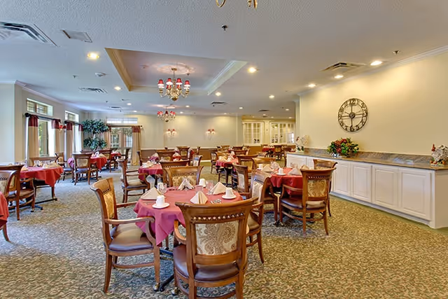 A spacious dining room with elegantly set tables featuring red tablecloths and decorative napkins. The room is well-lit with chandeliers and has a warm ambiance, complemented by plants and a wall clock.