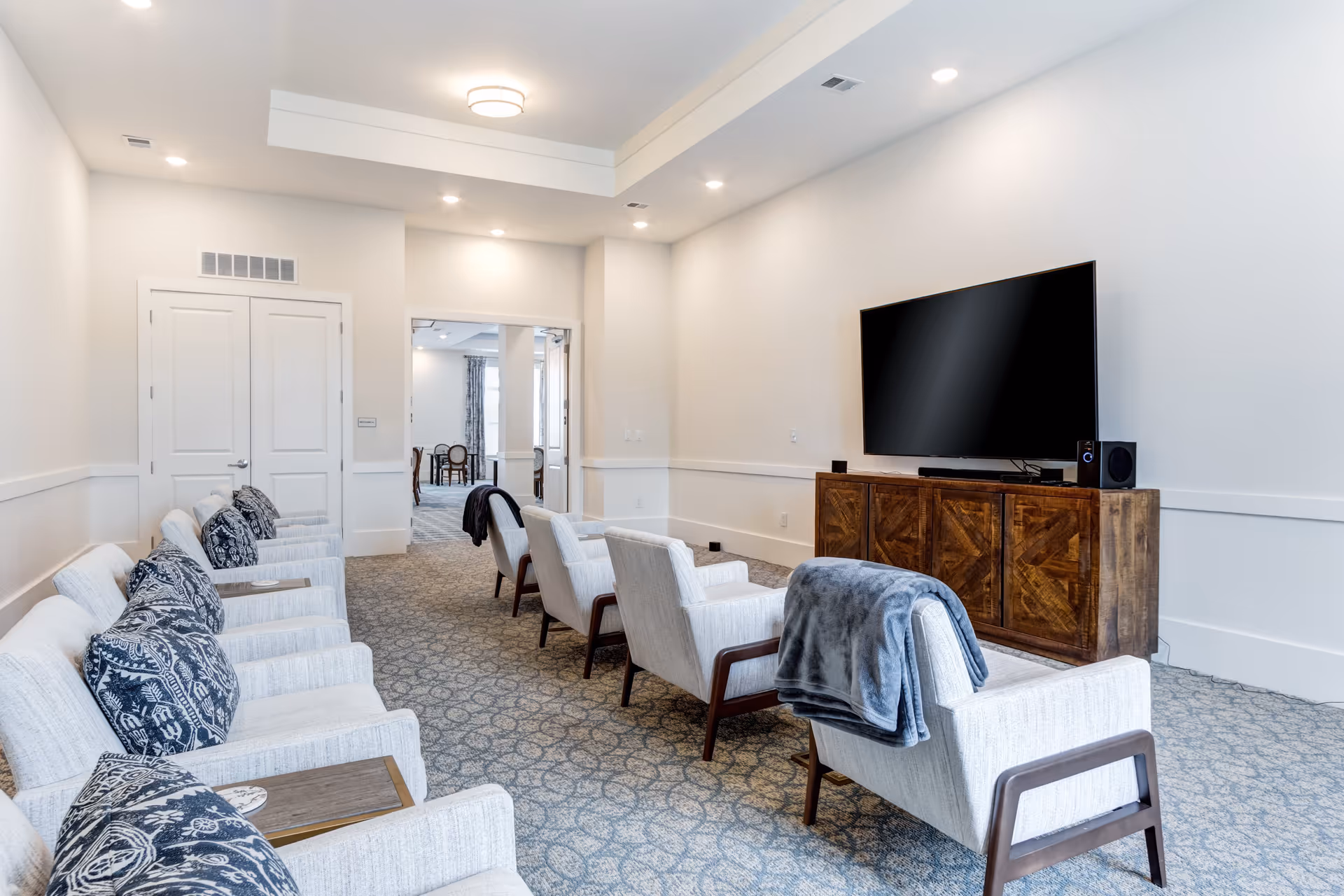 A bright and spacious lounge area with a row of light gray armchairs featuring dark wooden armrests and blue patterned cushions. A large flat-screen TV is mounted on a wooden cabinet against the wall. The room has white walls, recessed lighting, and a patterned carpet. In the background, there is an open doorway leading to another room with tables and chairs.