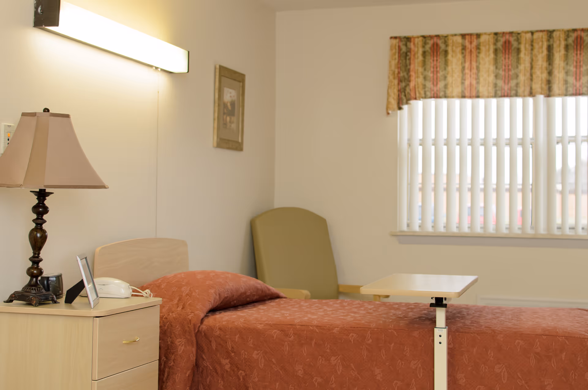 A simple bedroom in a senior living facility featuring a single bed with a rust-colored bedspread, a wooden nightstand with a lamp, a telephone, and a picture frame. There is a green armchair in the corner and a window with vertical blinds and a patterned valance.