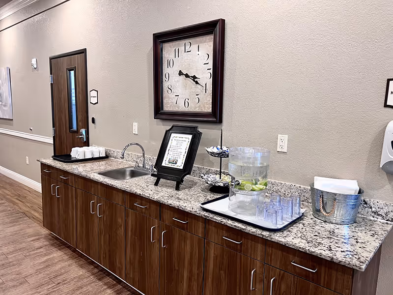 A countertop area in a senior living facility with a sink, a large wall clock, a framed sign, a tiered tray with wrapped items, a water dispenser with lime slices and plastic cups on a tray, and a metal bucket with napkins. The countertop is granite with wooden cabinets below, and the wall is beige with a wooden door and a hand sanitizer dispenser mounted on the wall.