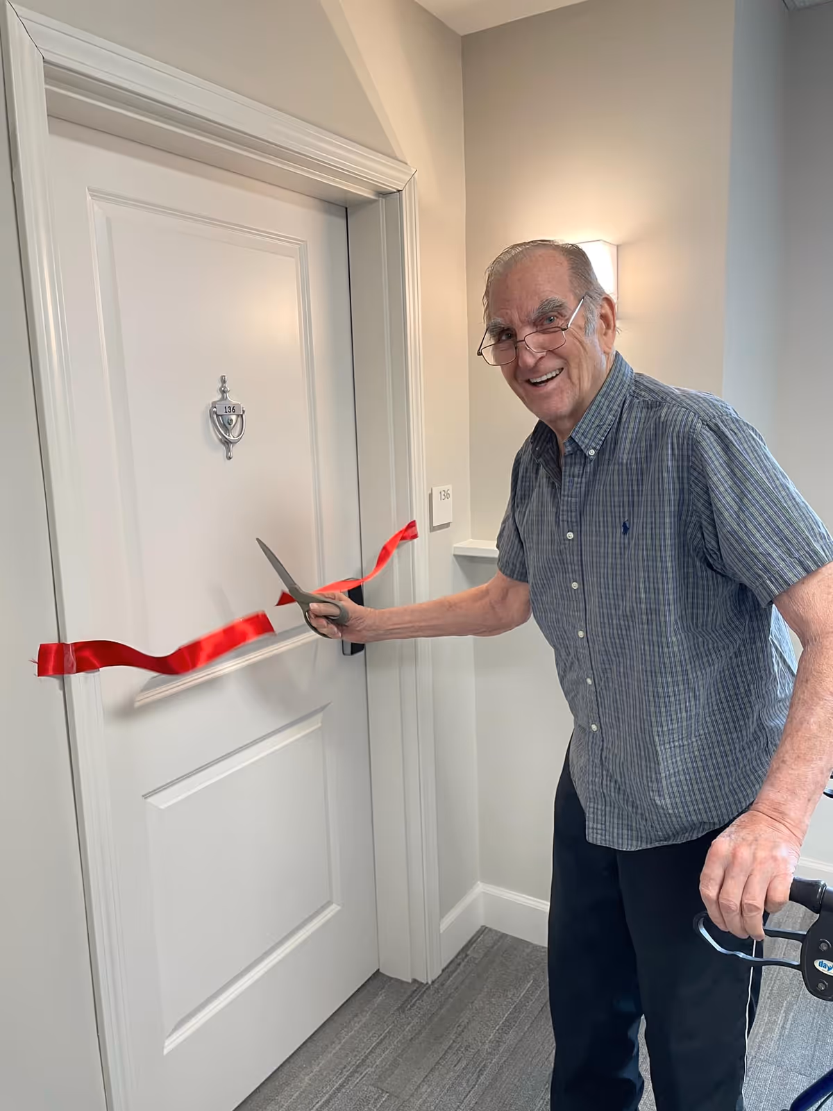 An elderly man with glasses and a plaid shirt is smiling while cutting a red ribbon stretched across a white door numbered 136 inside a hallway. He is holding a walker with one hand and large scissors with the other, appearing to celebrate an opening or special event.