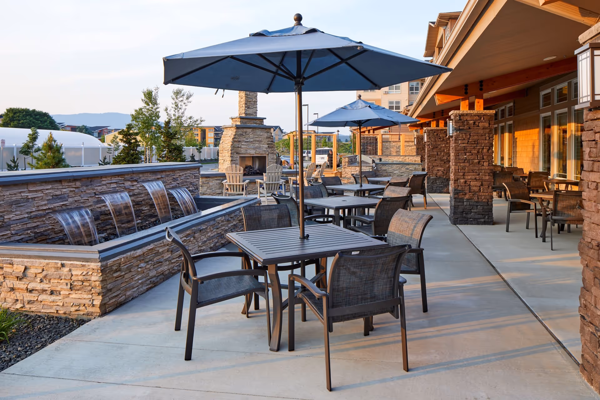 Outdoor patio area with multiple tables and chairs under blue umbrellas, stone pillars, a stone fireplace, and a water feature with cascading water, set against a backdrop of trees and distant buildings under a clear sky.