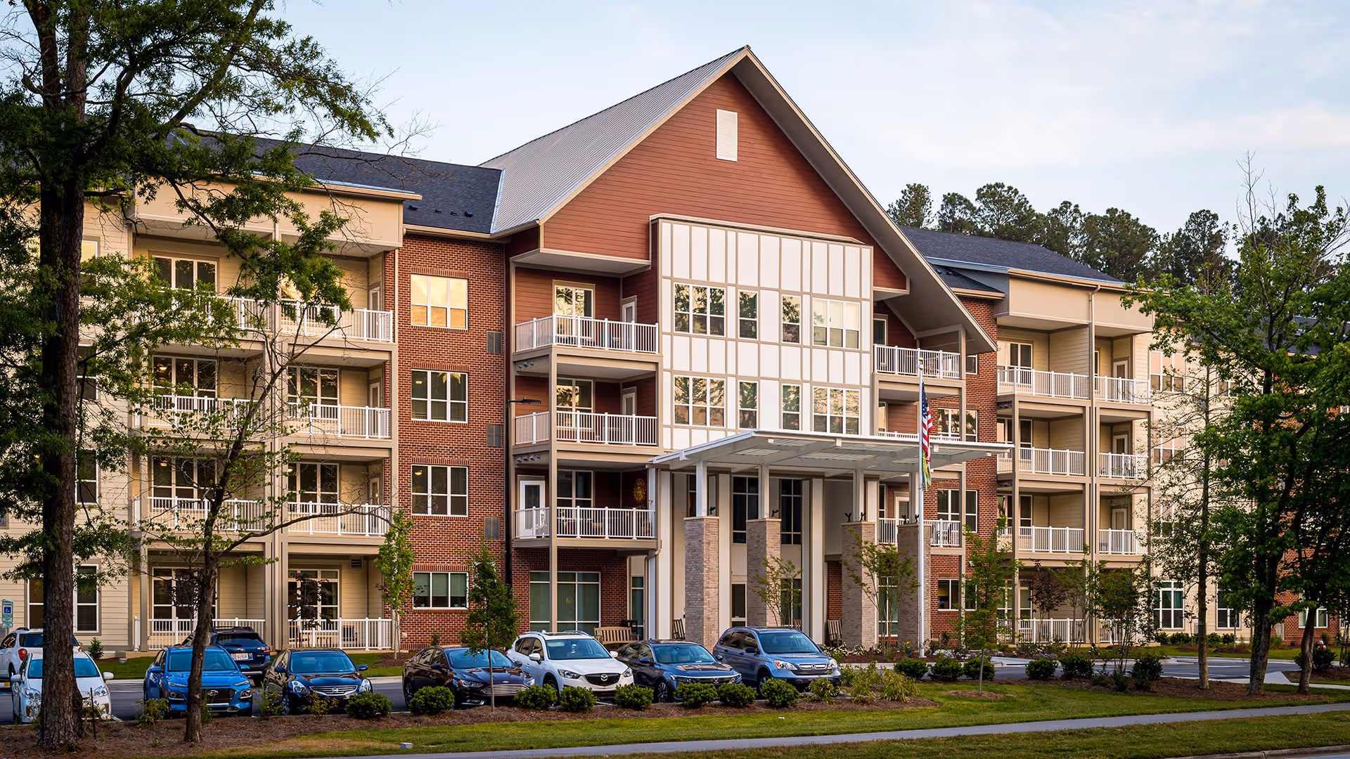 Exterior view of a multi-story senior living facility named Atria Cary, featuring a large entrance with a covered porch, balconies on each floor, surrounded by trees and parked cars in front.