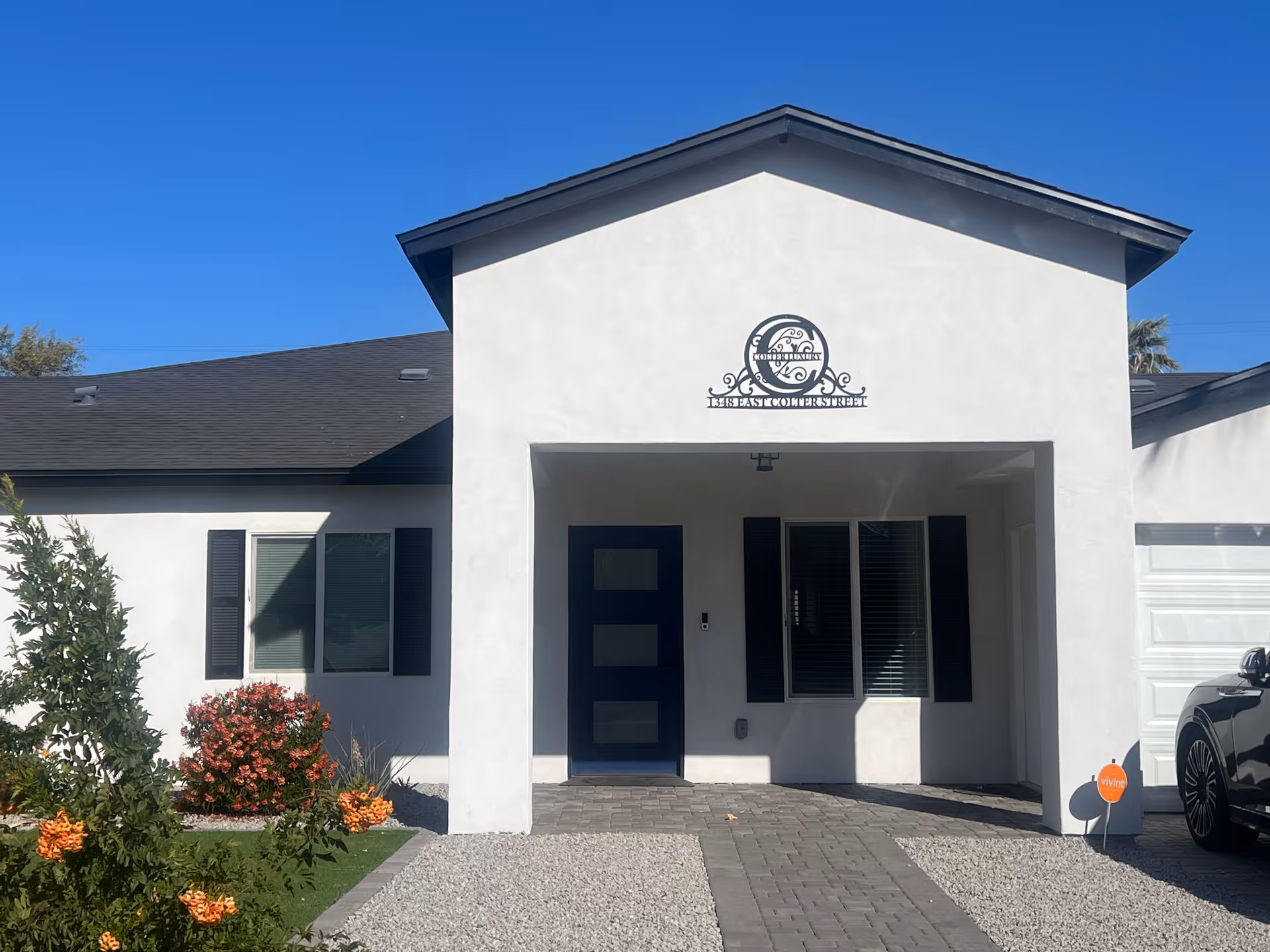 Front entrance of a white single-story building with a covered porch, dark blue door, shuttered windows, and a landscaped gravel driveway under a clear blue sky.