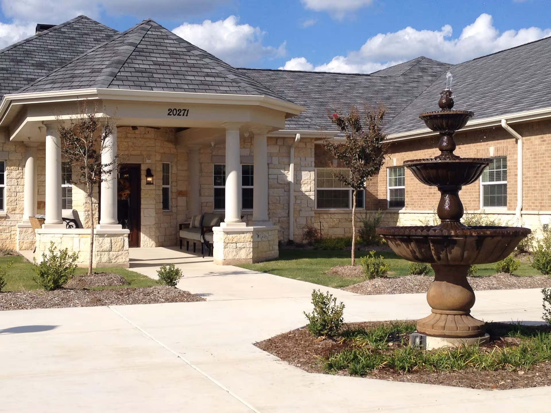 Exterior view of The Canyons of Stone Oak Memory Care facility showing a stone and brick building with a covered entrance supported by white columns, a concrete walkway, small landscaped bushes and trees, and a three-tiered water fountain in the foreground under a partly cloudy sky.