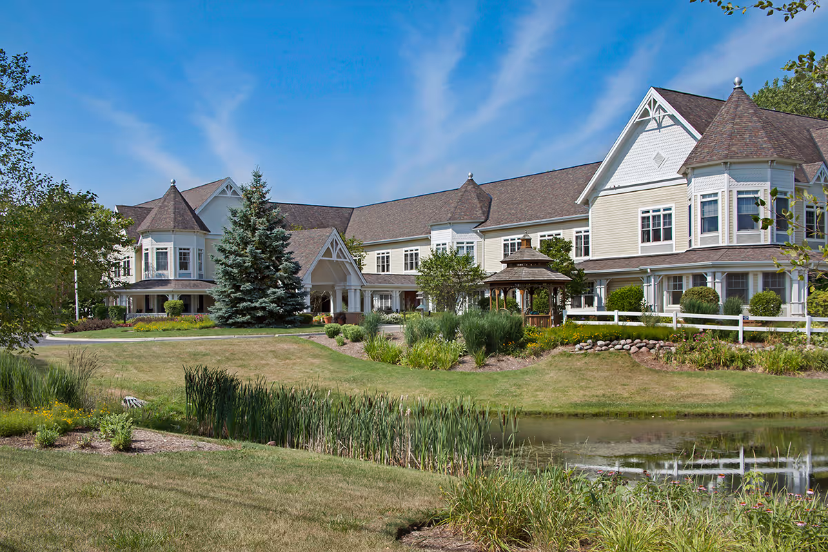 A large two-story senior living building with a gazebo, landscaped grounds, and a reflecting pond under a blue sky.