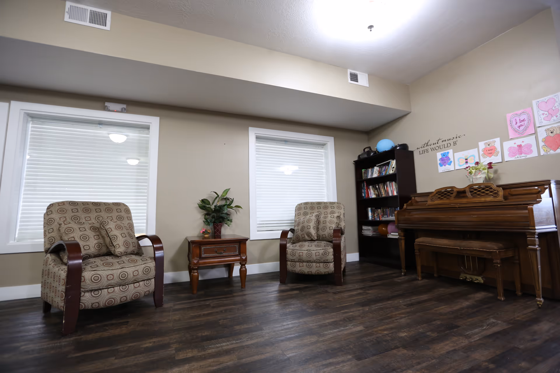 A cozy room with two patterned armchairs separated by a wooden side table with a plant on it, two windows with closed blinds, a dark wooden bookshelf filled with books, and a wooden piano with a bench. The wall above the piano has colorful drawings and a quote that reads 'without music LIFE WOULD B'. The floor is dark wood, and the walls are painted beige.