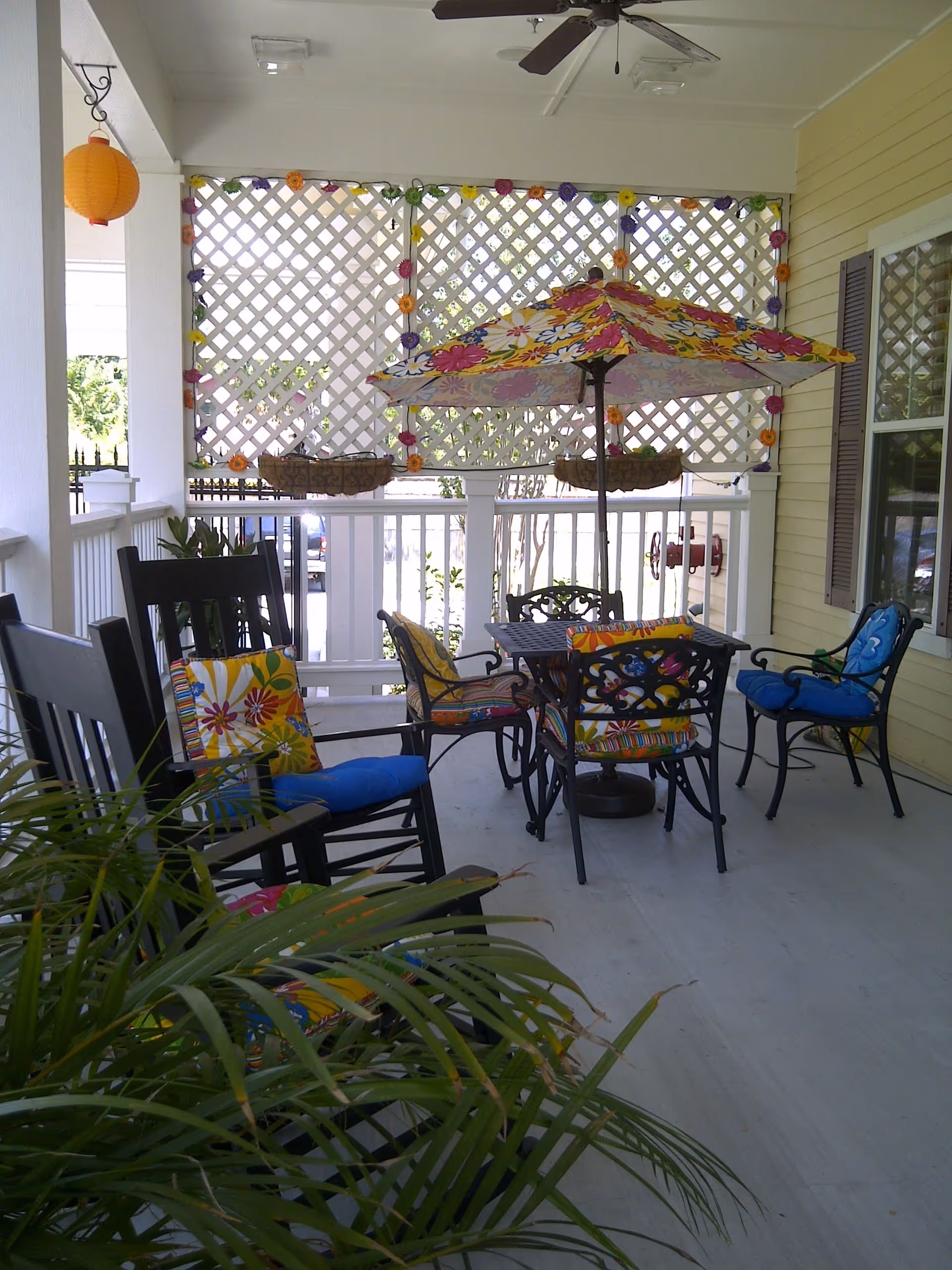 A covered outdoor patio area with a table and four chairs, each with colorful floral cushions. A large floral umbrella is positioned over the table. There are two black rocking chairs with blue and floral cushions on the left side. The patio has white railings and lattice panels decorated with small colorful flowers. A ceiling fan is mounted above, and a potted plant is visible in the foreground.
