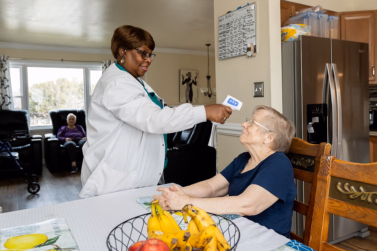 A caregiver uses a forehead thermometer to check an elderly woman's temperature at a dining table in a senior living facility.
