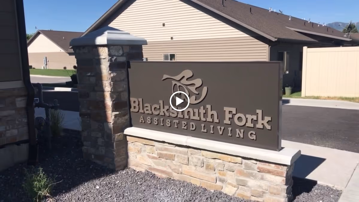 Stone and brick sign for Blacksmith Fork Assisted Living located outdoors near a driveway with residential buildings in the background under a clear blue sky.