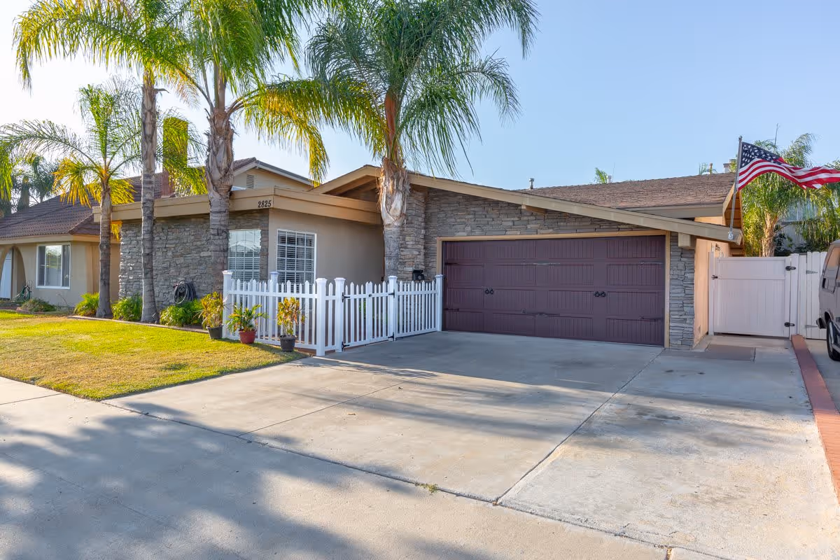 Exterior view of a single-story house with a stone facade and a brown garage door. The house has a small white picket fence with potted plants in front, several tall palm trees, and an American flag on a pole near the driveway. The driveway is concrete and the sky is clear and blue.