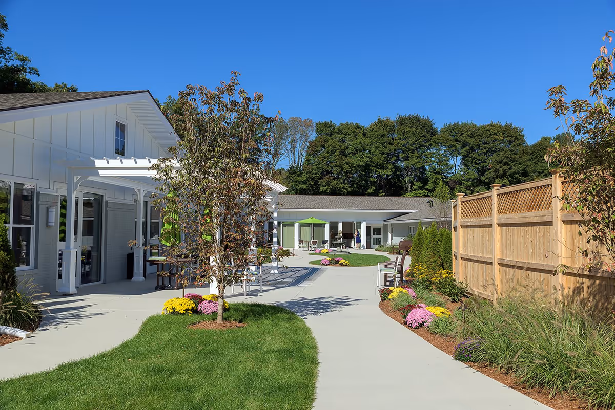 Outdoor courtyard area at Seasons of Danvers featuring a paved walkway, green grass, colorful flowers, small trees, wooden fence, and white buildings with large windows under a clear blue sky.