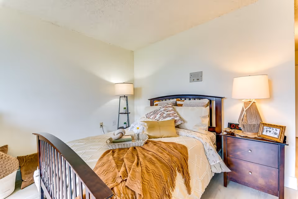 A cozy bedroom with a wooden bed frame, beige bedding, and multiple pillows. A brown throw blanket is draped over the bed. There is a small wooden nightstand with a lamp, a framed photo, and an alarm clock. A floor lamp with a small shelf stands in the corner. The walls are light-colored and the room has a warm, inviting atmosphere.