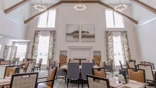 A spacious dining room with high vaulted ceilings and exposed wooden beams. The room features large windows with patterned curtains allowing natural light to fill the space. There are multiple dining tables with chairs arranged neatly, and a white fireplace with two framed artworks above it is centered on the far wall.