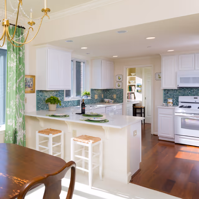 Bright kitchen with white cabinetry, a central breakfast bar with two stools, green mosaic backsplash and matching green curtains.