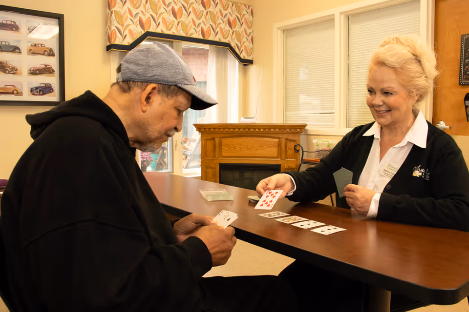 An elderly man and a woman sitting at a table playing cards in a cozy room with a fireplace and windows in the background. The woman is smiling and holding cards, while the man is looking at his cards. The room has warm lighting and framed pictures on the wall.