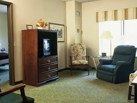 A cozy living room area with a floral upholstered armchair, a blue recliner, a wooden TV cabinet with a small television, a side table with a lamp, and a window with striped valance. A doorway leads to a bedroom with a bed visible.