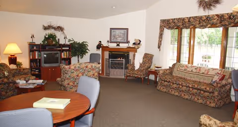 A cozy living room in a senior living facility featuring floral patterned sofas and armchairs, a wooden round table with chairs, a fireplace with decorative items on the mantel, a TV on a wooden stand, a large window with floral valance curtains, and a floor lamp providing warm lighting.