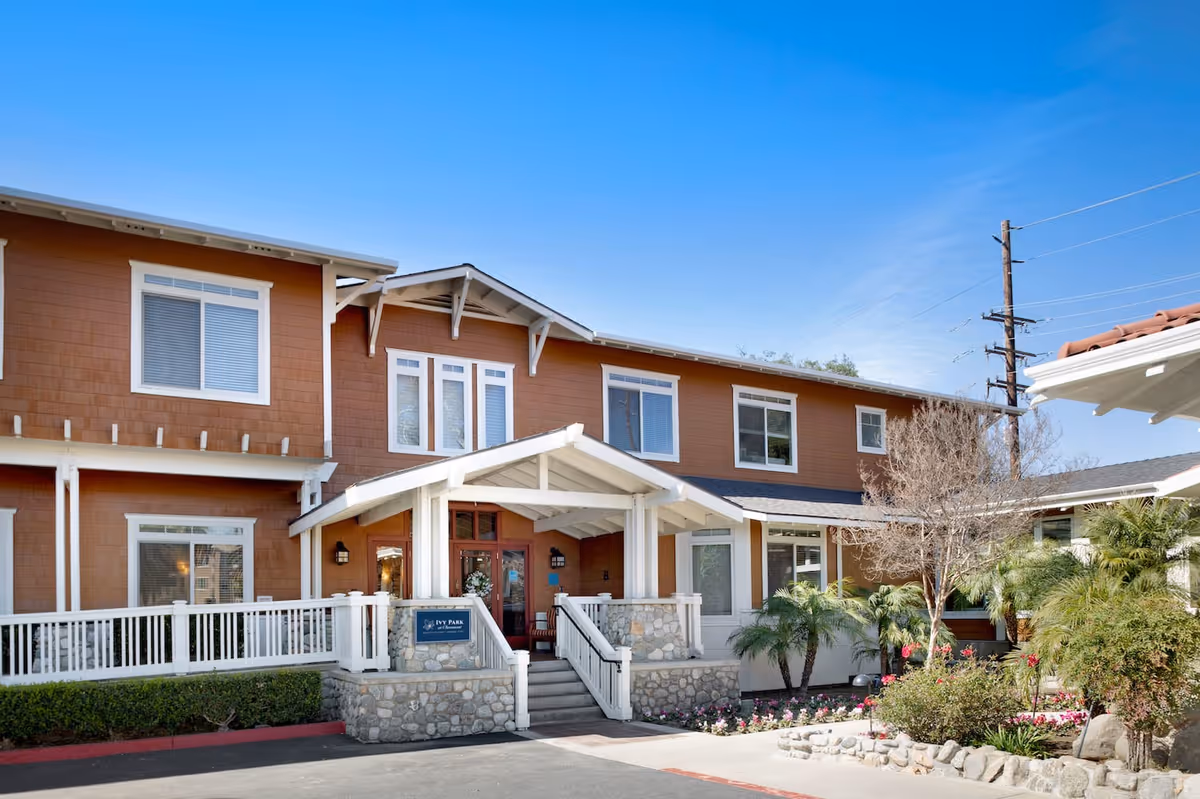 Exterior view of Ivy Park at Claremont, a two-story senior living facility with brown siding and white trim. The entrance features a covered porch with stone pillars and steps leading up to the front door. There are landscaped bushes, palm trees, and flowers around the building under a clear blue sky.