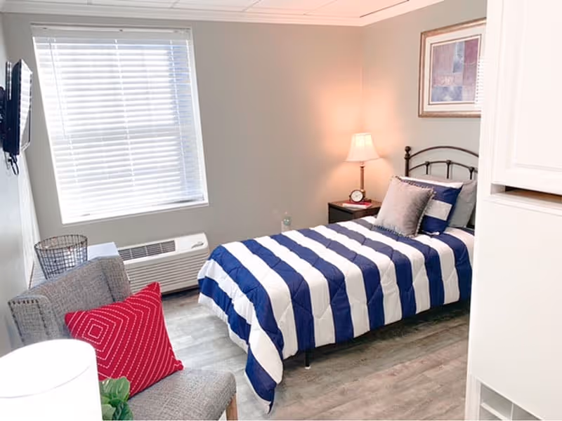 A small, neatly arranged bedroom with a single bed featuring a blue and white striped comforter and multiple pillows. There is a gray armchair with a red pillow, a nightstand with a lamp and clock, a wall-mounted TV, and a window with blinds letting in natural light.