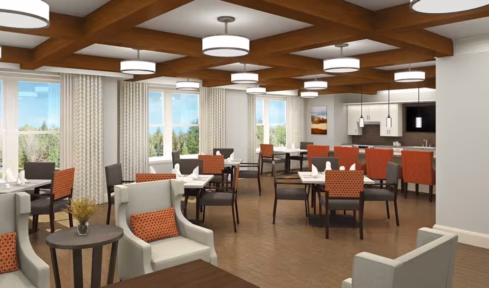 A bright and spacious dining room with multiple tables and chairs arranged neatly. The room features large windows with curtains allowing natural light to fill the space. There are modern ceiling lights with wooden beams on the ceiling. The dining area includes a kitchen counter with bar stools and pendant lights hanging above it. The chairs have a mix of solid and patterned upholstery in shades of gray and orange.