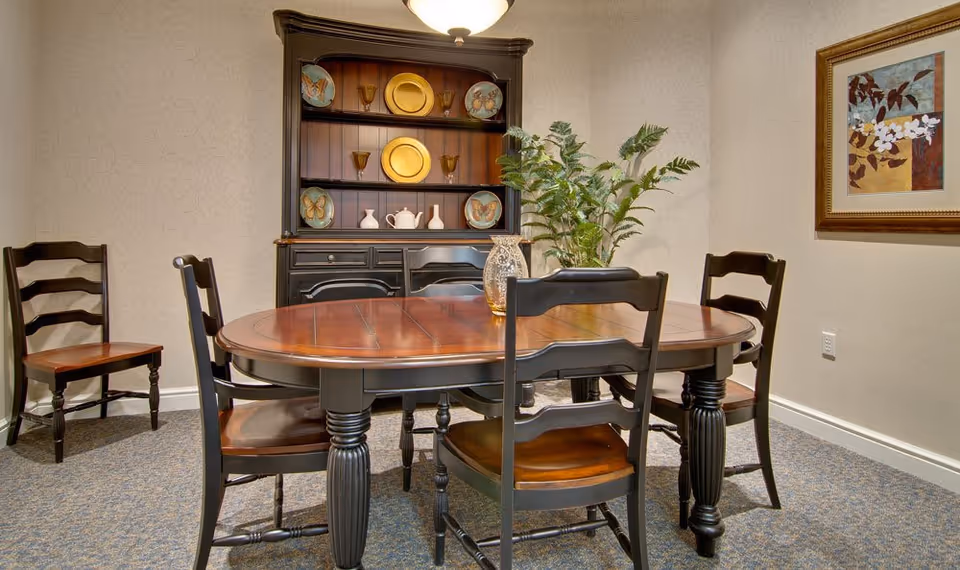 Wooden dining table with chairs in a cozy dining room featuring a dark hutch, potted plant, and framed artwork.