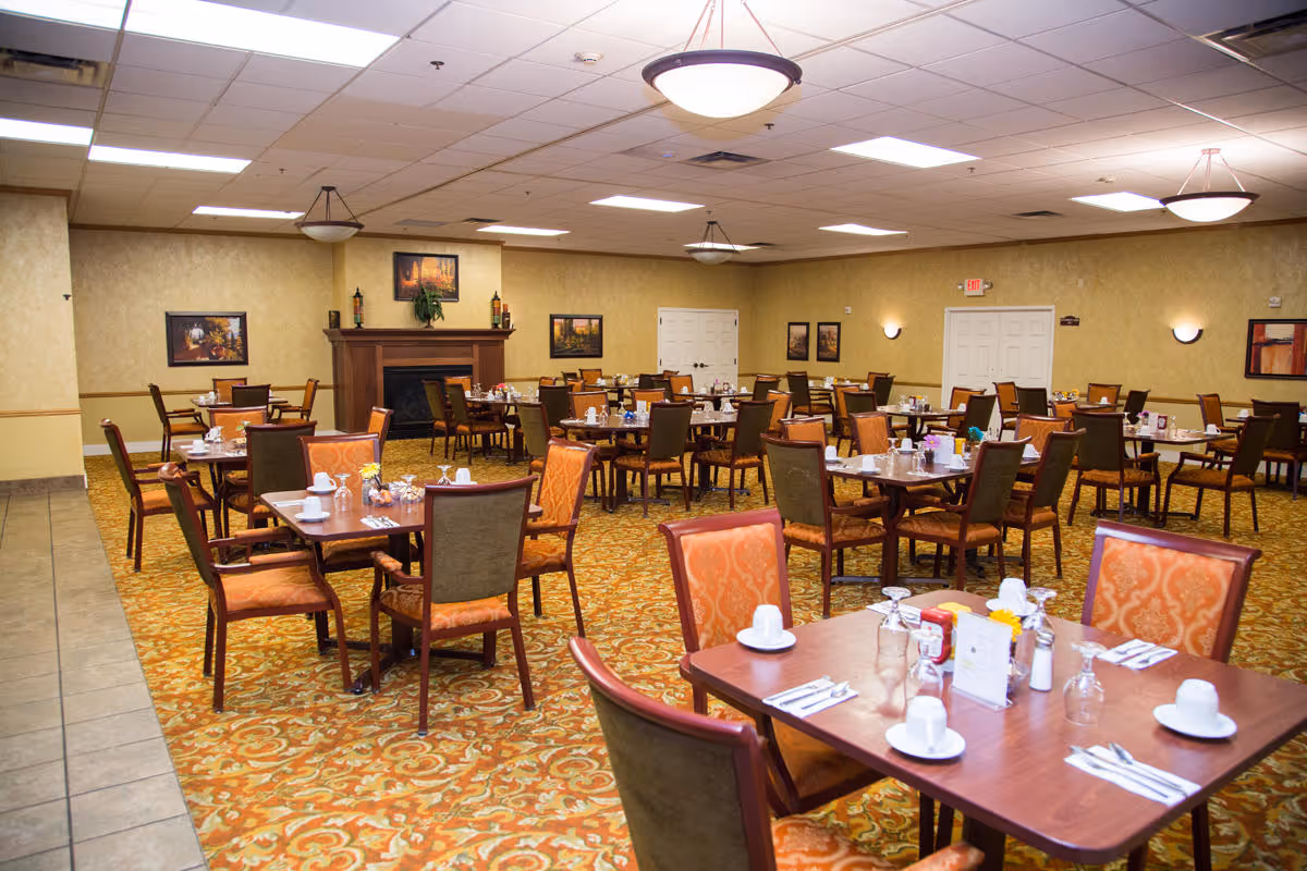 A spacious dining room with multiple wooden tables and chairs arranged neatly. Each table is set with white cups, glasses, utensils, and condiments. The room has warm yellow walls, a patterned carpet, ceiling lights, and framed artwork on the walls. There is a fireplace in the background.
