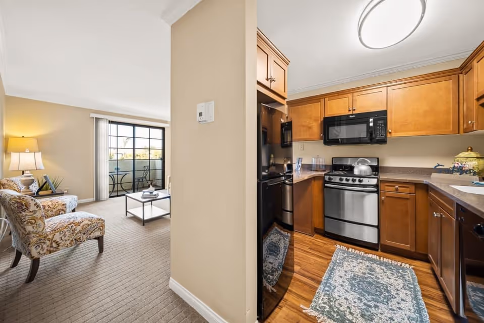 Interior view showing a cozy living room with patterned armchairs, a coffee table, a lamp, and a sliding glass door leading to a balcony. Adjacent to the living room is a kitchen with wooden cabinets, stainless steel appliances including a refrigerator, stove, microwave, and dishwasher, and a decorative rug on the wooden floor.