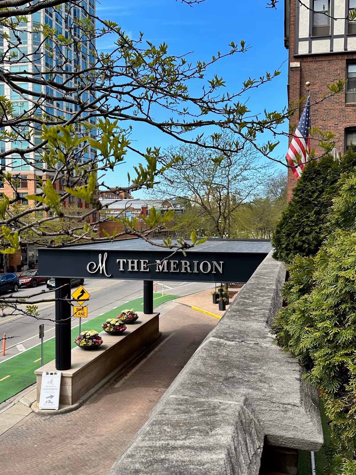 View of the entrance canopy of The Merion building with its name displayed on the front. The scene includes a sidewalk with flower pots, a bike lane on the street, trees with green leaves, and part of a brick building with an American flag.