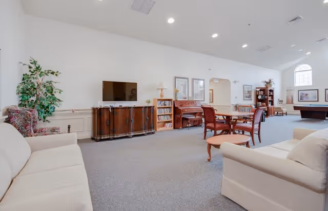 A spacious living room area in a senior living facility with beige sofas, a wooden TV stand with a flat-screen TV, a piano, a round wooden table with chairs, bookshelves, and a pool table in the background. The room has high ceilings with recessed lighting and large windows allowing natural light.