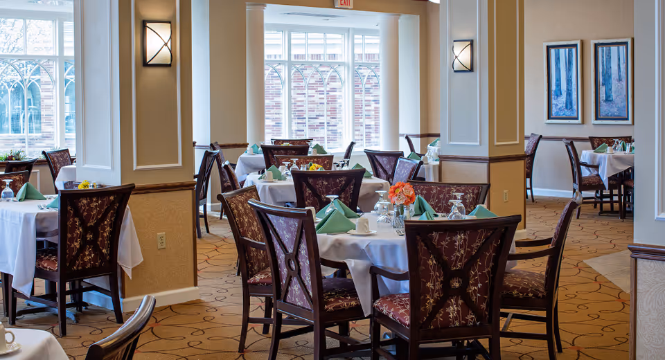 Dining room with round tables set with white tablecloths, green napkins, floral centerpieces, and upholstered chairs by large windows.