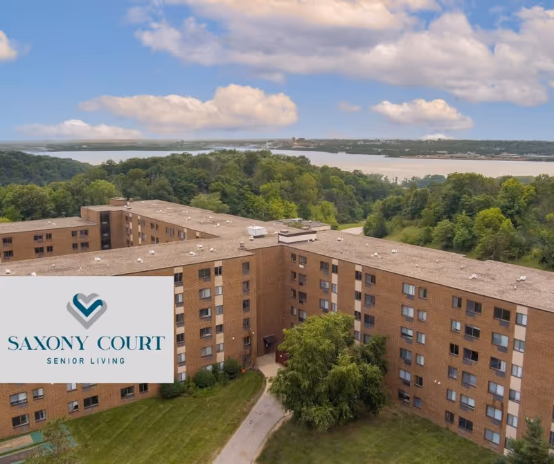 Aerial view of Saxony Court Senior Living, a large multi-story brick building surrounded by green trees and grass, with a river and forested area in the background under a partly cloudy sky.