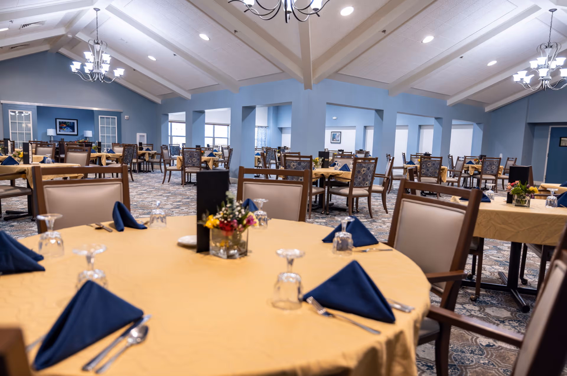 A spacious dining room with multiple tables covered in yellow tablecloths, each set with blue folded napkins, upside-down glasses, and silverware. The room features blue walls, patterned carpet, and chandeliers hanging from a vaulted ceiling. There are floral centerpieces on the tables and framed artwork on the walls.