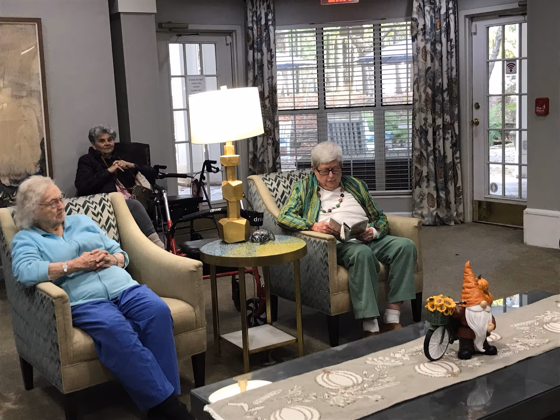 Three elderly women sitting in a living room area of a senior living facility. Two women are seated in armchairs, one reading a book and the other resting with hands clasped. The third woman is seated in a wheelchair near the back. The room has large windows with blinds and floral curtains, a table with a lamp, and a decorative gnome figurine on a coffee table.
