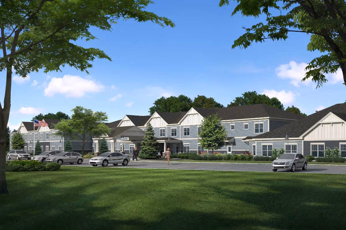 Front exterior view of a two-story senior living facility with a landscaped lawn, parked cars, and an American flag.