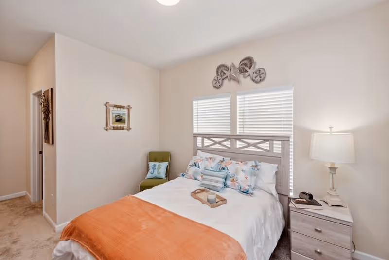 A cozy bedroom with a bed featuring a white comforter, floral and striped pillows, and an orange throw blanket. There is a wooden nightstand with a lamp, a phone, and a book. Two windows with white blinds are behind the bed, and above them are decorative sea shells. A green chair with a pillow is placed in the corner next to a framed picture on the wall.