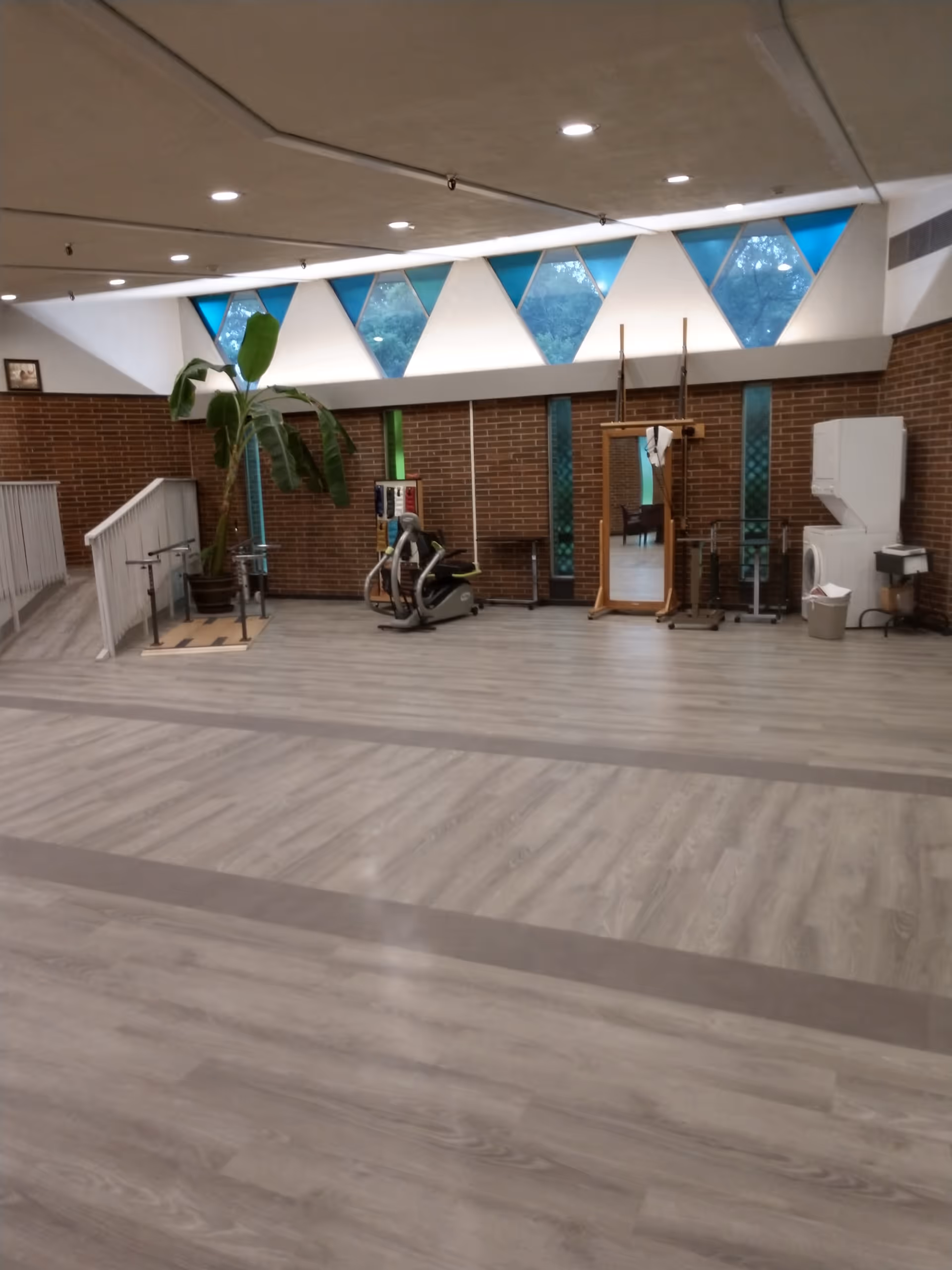 A spacious room with light wood flooring and brick walls, featuring exercise equipment including a stationary bike and parallel bars. There is a large mirror on a wooden stand, a potted plant, and triangular windows near the ceiling letting in natural light.