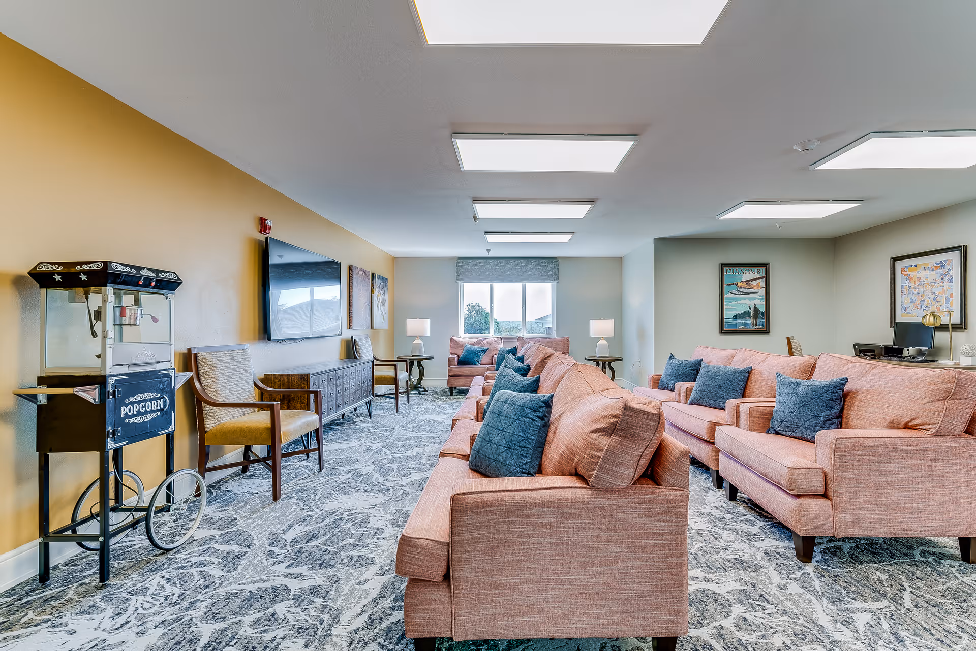 A bright senior living facility common area with multiple pink sofas adorned with blue cushions, a patterned carpet, a popcorn machine on the left, a flat-screen TV mounted on a yellow wall, and framed artwork on the walls. The room is well-lit with ceiling lights and natural light coming from a window at the far end.