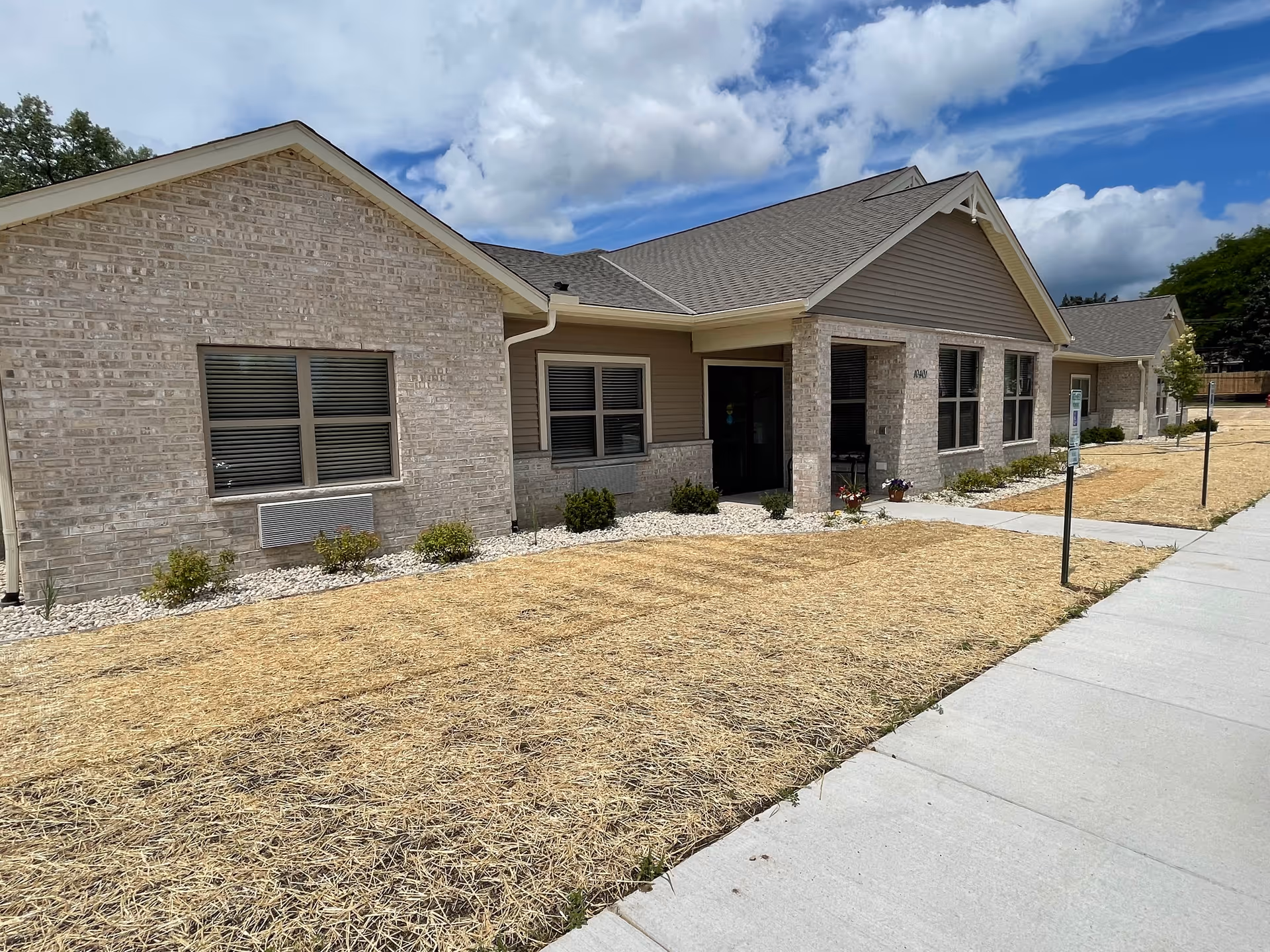 Single-story brick and siding assisted living building facade with windows, entrance, sidewalk, and straw-covered front yard under a partly cloudy sky.